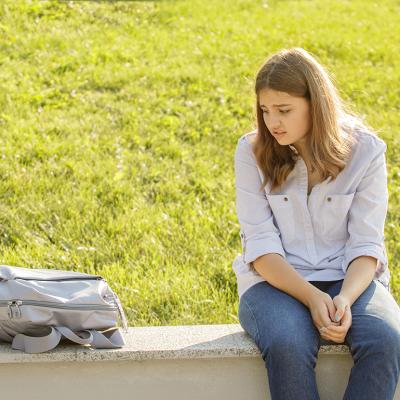An upset teenage girl sitting on a curb and staring at her backpack for school.