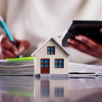 A small figure of a home with a real estate professional calculating finances in the background.