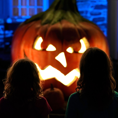 Two young girls standing in front of a giant lit up jack-o'-lantern at night.