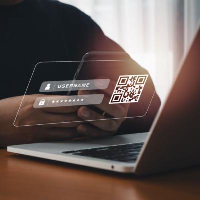 A person holding a smartphone in front of a laptop with an overlaid image of a log in screen and QR code. 
