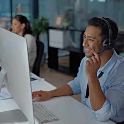 A young man smiling, using a headset while looking at the computer in a modern office.