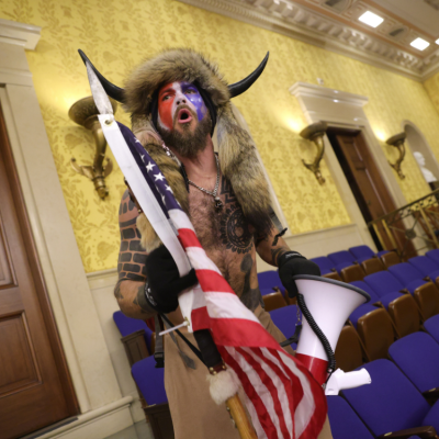 During the Capitol attack, Jacob Chansley, shirtless and wearing a horned helmet and a fur hat with his face painted in red, white, and blue, yells while holding an American flag with a spear attached top the end and a megaphone. 