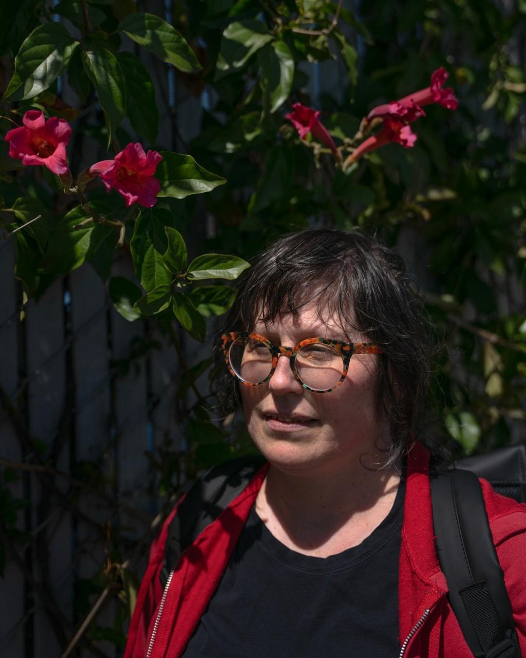 Julie Crossman in the sun in front of pink trumpet-shaped flowers that are in the shade, in Oakland on April 8, 2025.