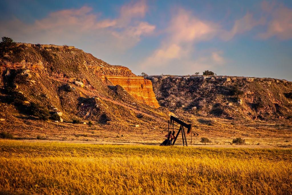 Oil well pump jack in red earth western Oklahoma at golden hour with early sunset sky and bluffs in background.