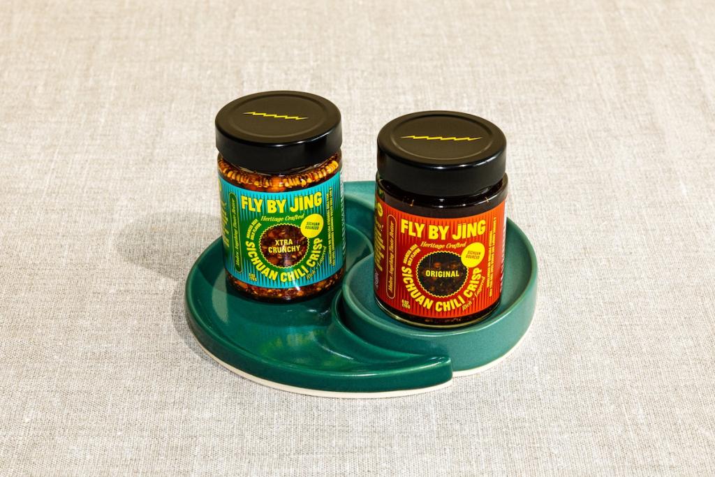Two jars of Fly By Jing chili crisp sitting on a green yin-yang shaped ceramic dish.