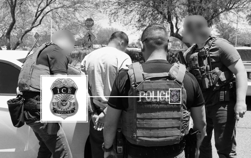 Black and white photo of group of four U.S. Immigration and Customs Enforcement (ICE) officers, wearing tactical vests and armed with weapons, detain a man in a white shirt with his hands cuffed behind his back next to a car. Enlarged inset shows detail of ICE patch on vest.