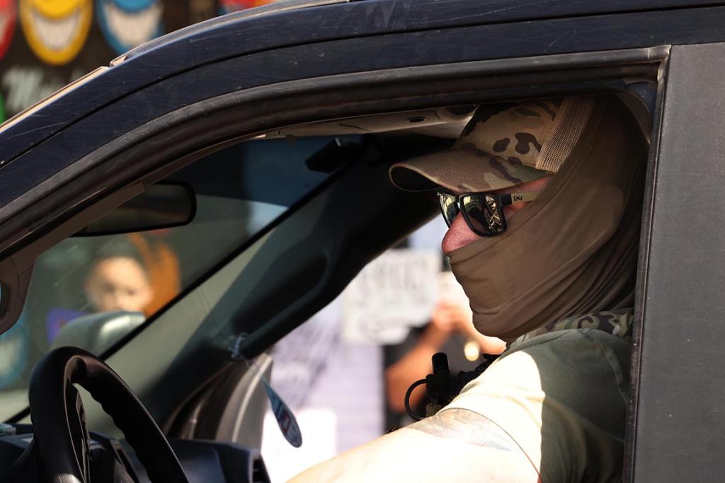 Closeup of a masked federal agent wearing sunglasses and a hat seen through his drivers' side window, planning his escape through an angry crowd after an immigrant raid on Atlantic Boulevard in the city of Bell
