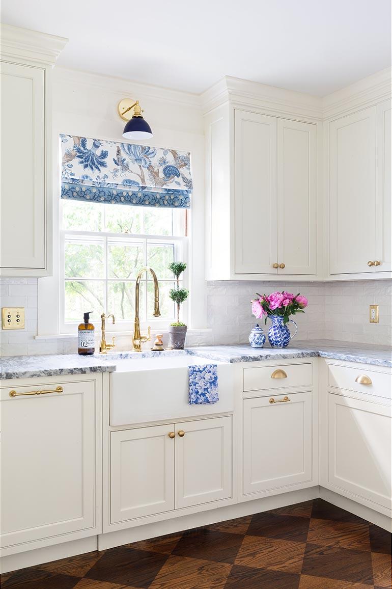 White cabinetry and a golden faucet with a window curtain that is blue and white like the wallpaper.