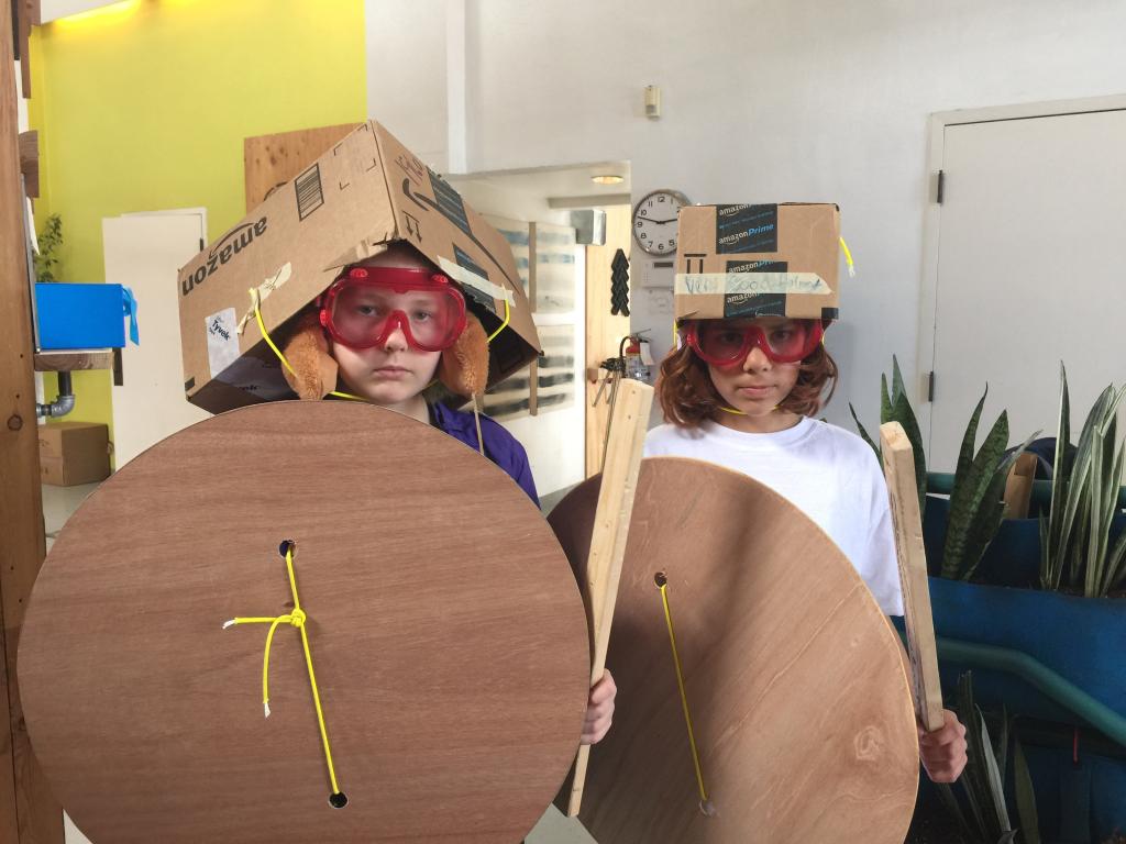 Two Brightworks students pose with their make-shift armor, shield, and swords made from wood and cardboard boxes.
