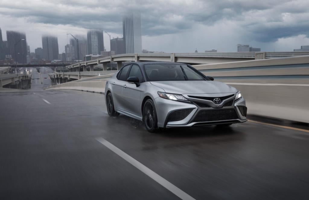Silver 2024 Toyota Camry driving on road with clouds and skyscrapers in the background.