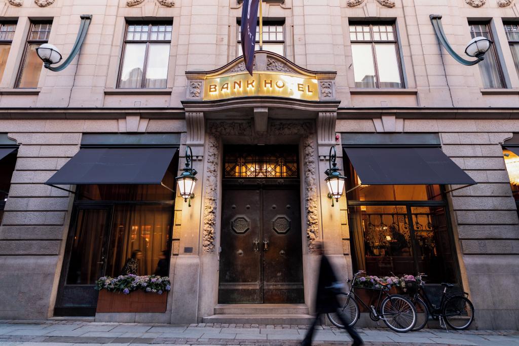 View of the entrance to the Bank Hotel with blurred person walking by and bicycles parked in front, the two imposing metal doors are apparent that it was historically a bank building.