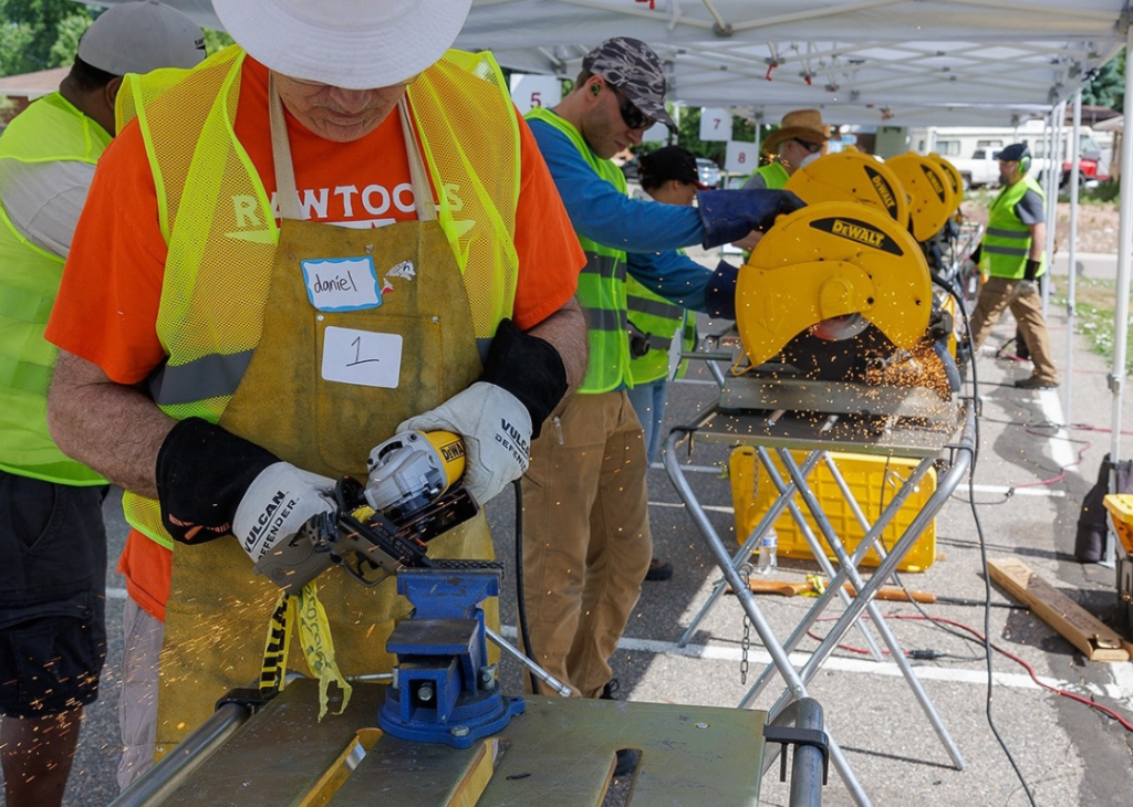 A row of people wearing safety vests and goggles using yellow DeWALT table saws which are throwing up sparks under EZ-up tents at a RAWtools live event. 