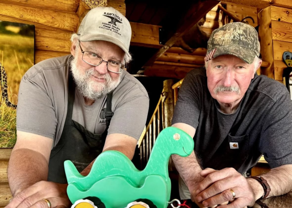 Two RAWtools blacksmiths who look like older men, one with a beard and the other with a mustache, pose with a with a green toy dinosaur made from parts of a disarmed gun. 