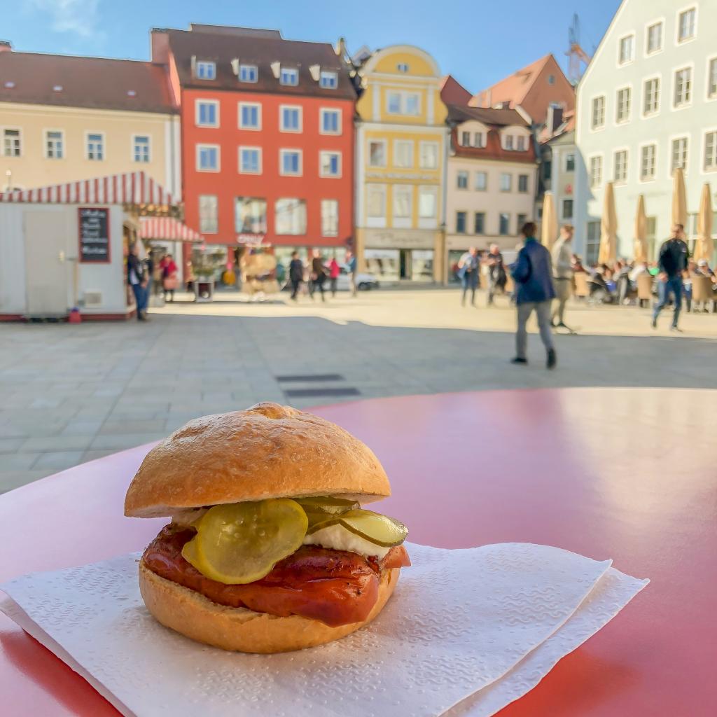 A Knacker, a one-of-a-kind Bavarian sausage served on a bun on a napkin on a red table in front of picturesque German buildings along the Danube River in eastern Bavaria.