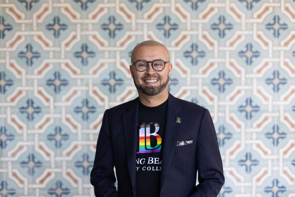 Mike Muñoz stands for a portrait in front of a tiled wall, wearing an LBCC tshirt with a rainbow logo underneath a sportcoat. He is a first-generation college student who now serves as superintendent and president of Long Beach City College and works to support students from traditionally disadvantaged backgrounds. 