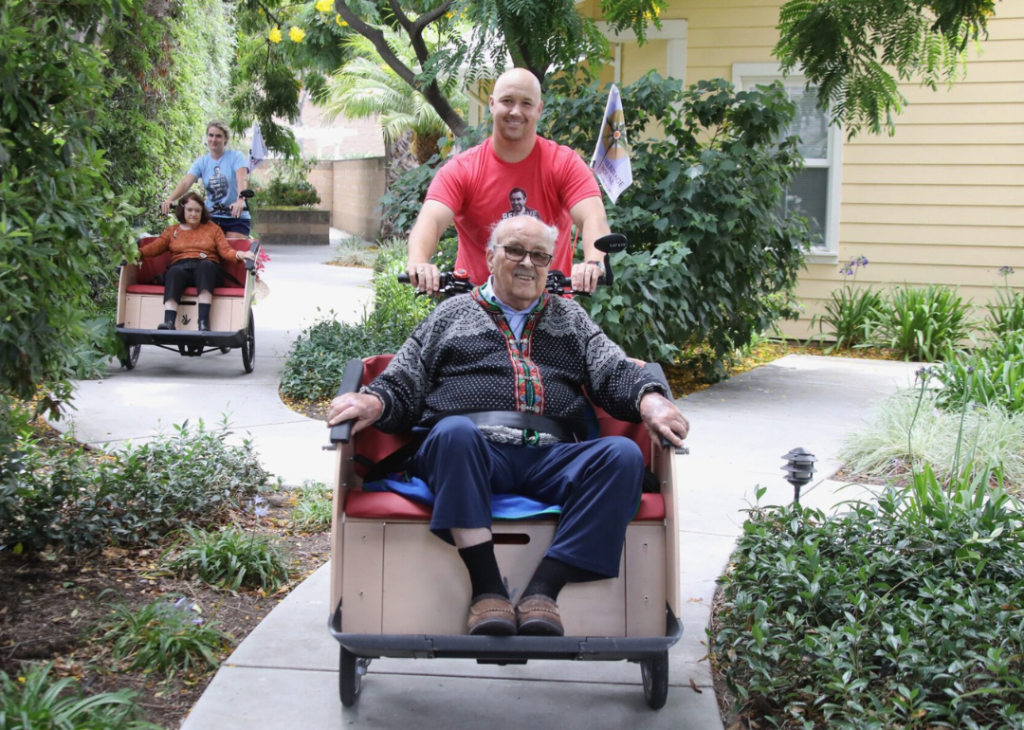 Two people from the volunteer team on trishaws, set up with a passenger sitting in front that can be pushed by a rider on a bicycle behind. 