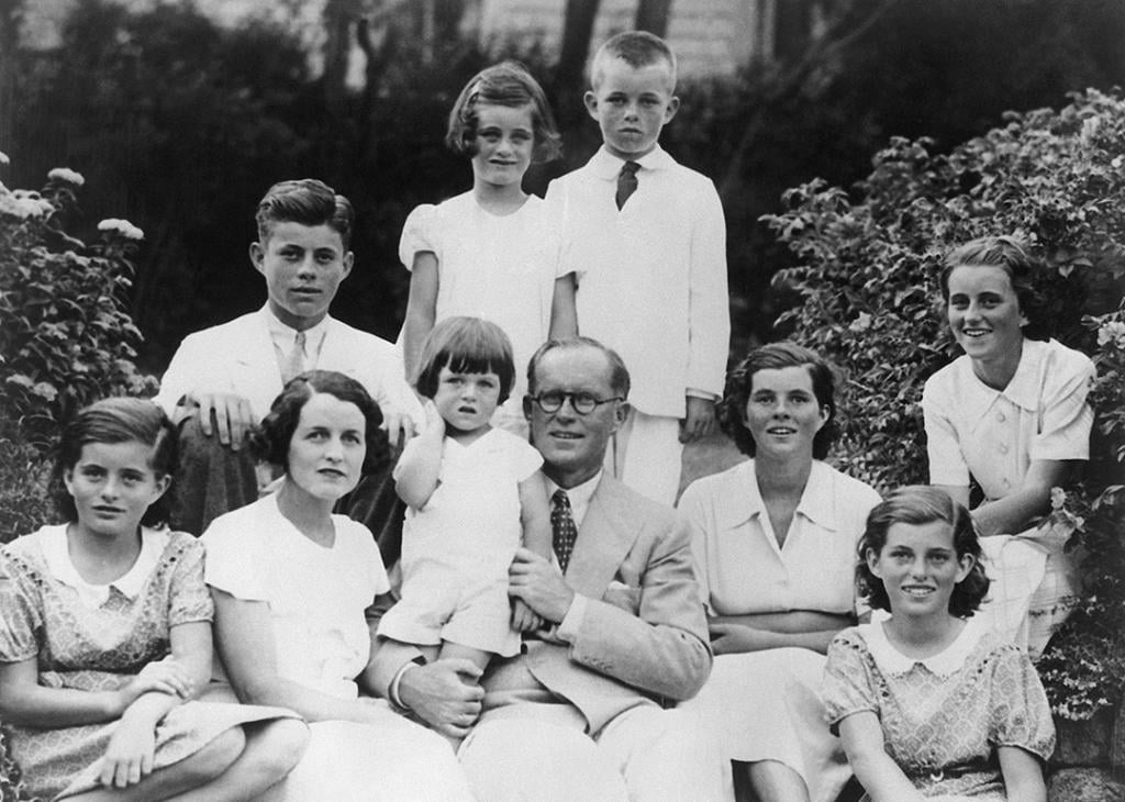 A black and white family portrait of the Kennedys. From left to right in the front row are Patricia, Rose and Joseph Kennedy, with baby Edward, Rosemary, Eunice, and Kathleen. The rear row is John, Jeanne, and Robert.