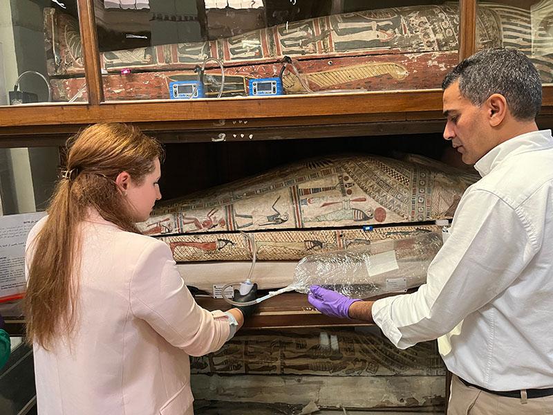 Two scientists wearing gloves and holding a device with tubing and a bag analyizing air next to mummies at the Egyptian Museum in Cairo so they can recreate their scent — and shed light on the embalming ingredients used to preserve them.