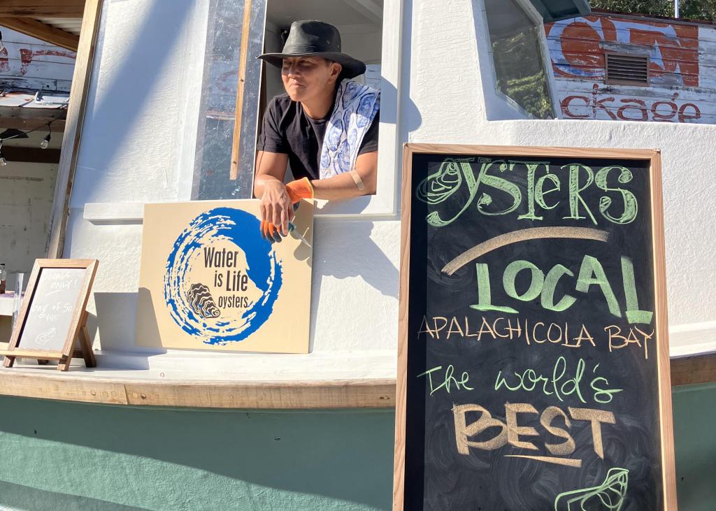 Kung Li, wearing a black hat, leans out the window of the old oyster boat that Li and business partner Xochitl Bervera repurposed to sell oysters directly to the community. Signs advertising the business are displayed in front.