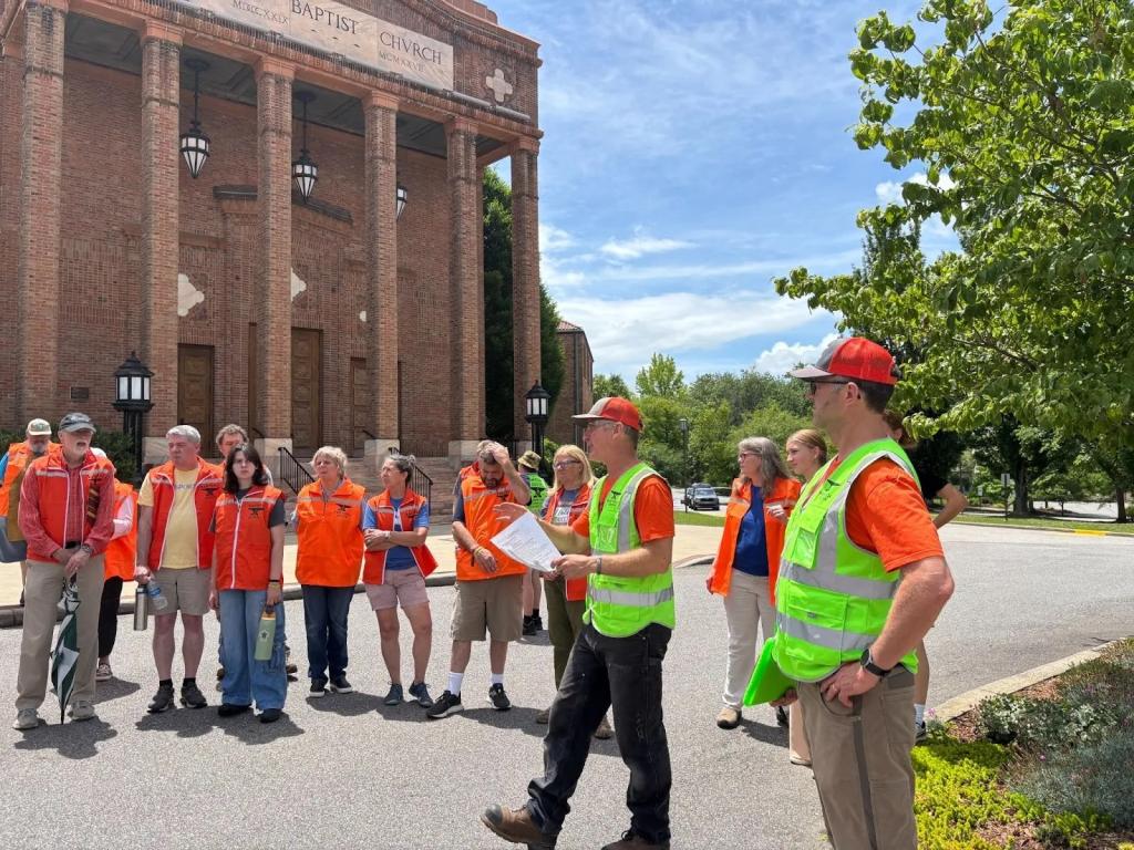 Utz and Wilson wearing high-viz safety vests, give instructions to a row of volunteers wearing orange at RAWtools South’s Guns to Gardens: Safe Disposal Event in Asheville, North Carolina.