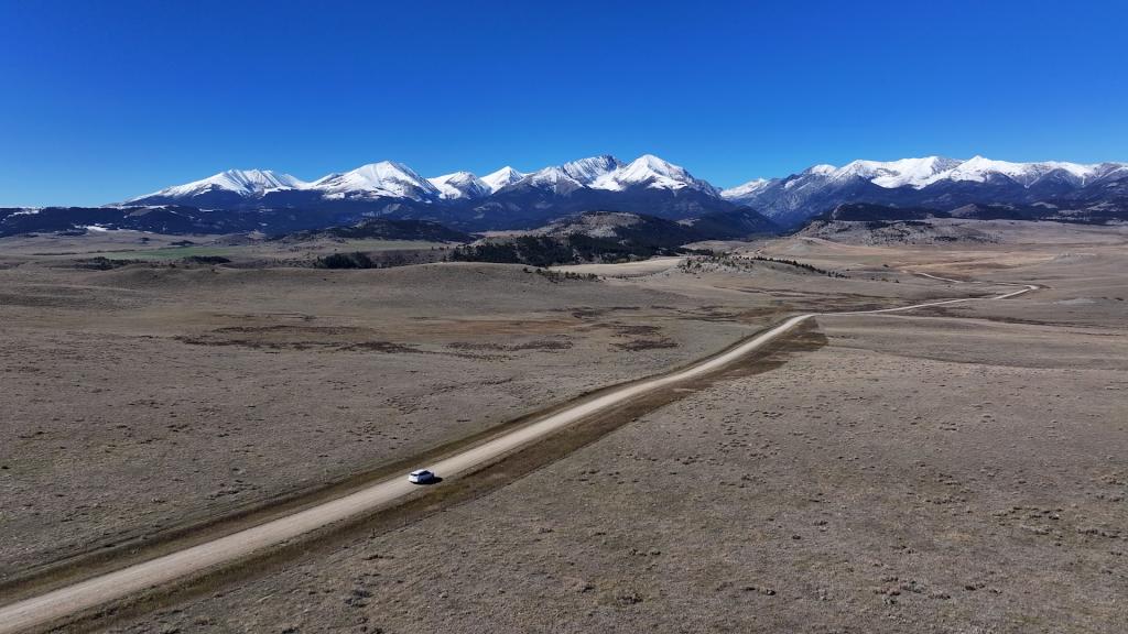 Aerial photo of single car driving on a long dirt road heading toward a mountain range, the Crazies. They resemble a mountain fortress — an island of jagged peaks rising more than 7,000 feet above the surrounding high plains, complete with secluded river valleys and alpine lakes.