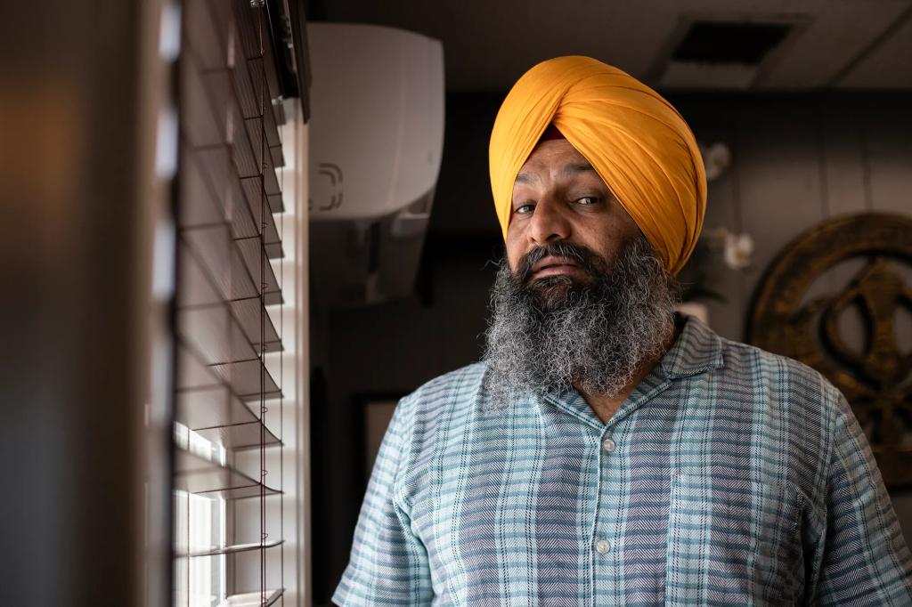 Harsimran Singh, who is president of the American Kabaddi Federation has a graying beard and a bright orange turban and he stands indoors beside a window with horizontal blinds. Soft light from the window falls across his face, creating gentle shadows. He wears a short-sleeve patterned button-up shirt and look toward the camera with a calm, steady expression. In the background, a circular wall decoration and parts of the room’s interior are visible.