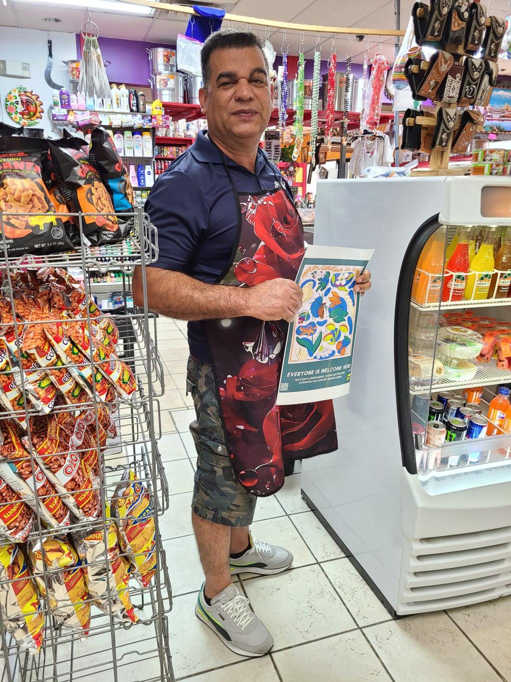 Man wearing a red floral apron holds a poster in a small standing between snack and drink displays. Resources for 4th Amendment Workplaces include a workplace guide, organizing toolkit, posters signaling opposition to unconstitutional search and seizures, employee handouts and tips for designating private employee areas.
