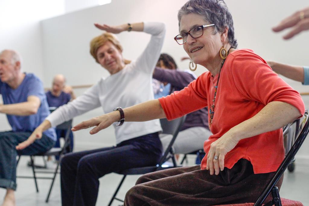 A woman in the Dance for PD program wearing an orange shirt dances from a chair in the foreground, while others are visible behind her doing the same.