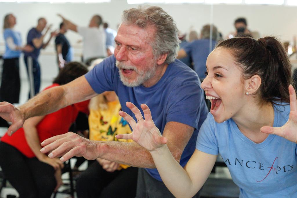 Two people wearing blue shirts, a young woman on the right next to an older man, make expressive faces with their arms raised in a Dance for PD program.