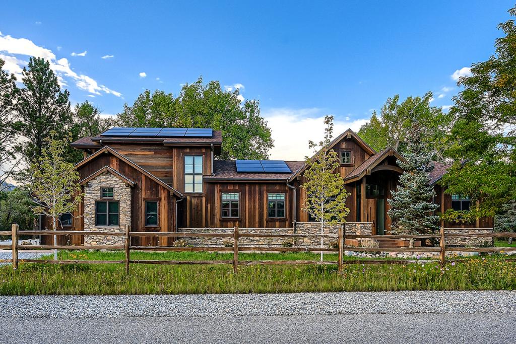 View of exterior of a house that is made with wooden and stone materials and has eco-friendly solar panels and native landscaping with a mixture of evergreen and deciduous trees planted in the yard.
