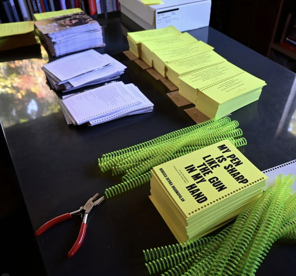 A small book called "My Pen is Sharp Like the Gun in My Hand" in the process of being bound with neon coils.