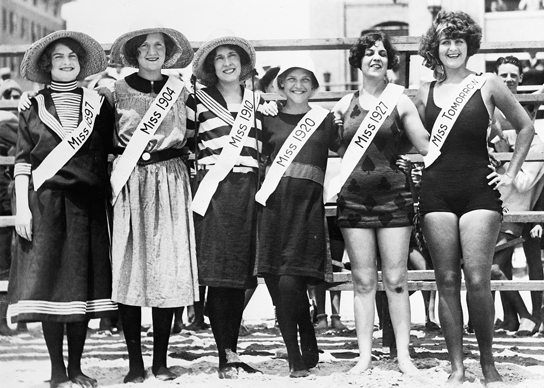 Six women pose wearing different swimsuits, with each wearing a sash that says "Miss" and the year the swimsuit was popular, including 1897, 1904, 1910, 1920, 1927, and tomorrow.