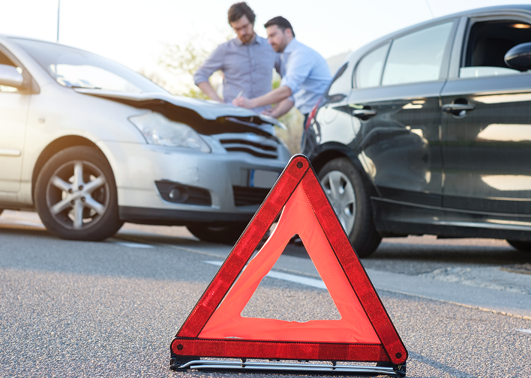 Two men standing by car crash with red emergency triangle on road.