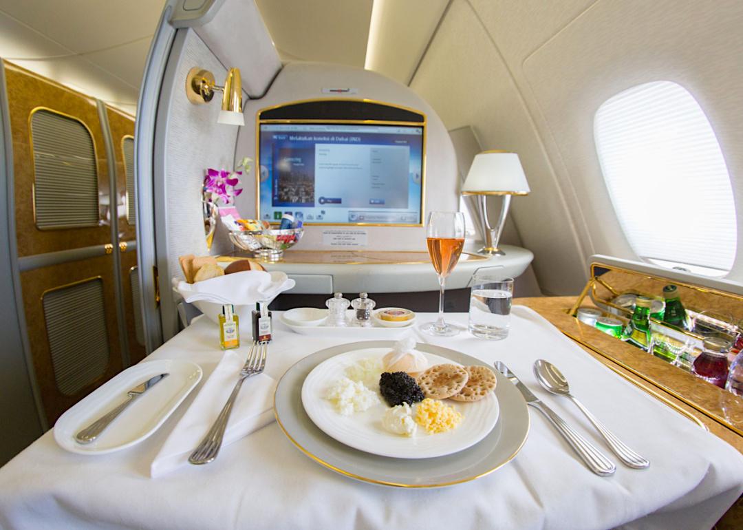 Emirates airlines first class suite with a large screen, a mini bar, and a table set with a meal and a drink in a glass flute.