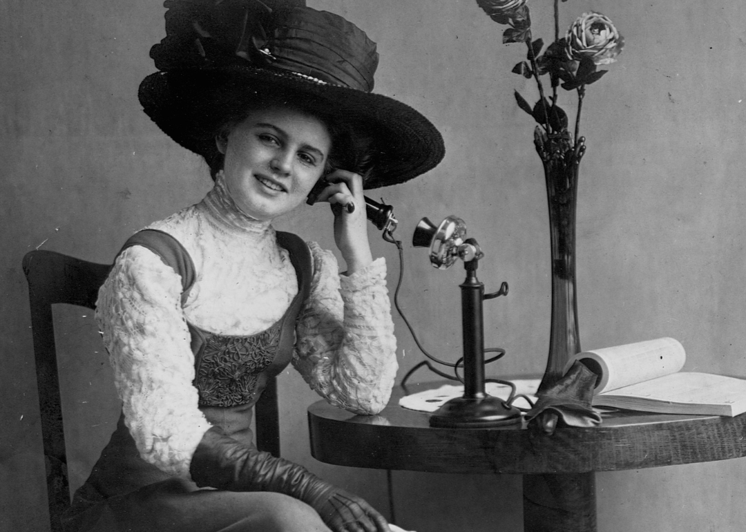 Woman with hat seated at table talking on early standing telephone.