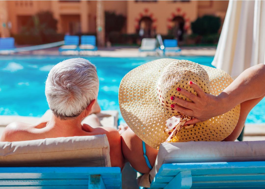Retired senior couple relaxing by swimming pool on chaise lounges.