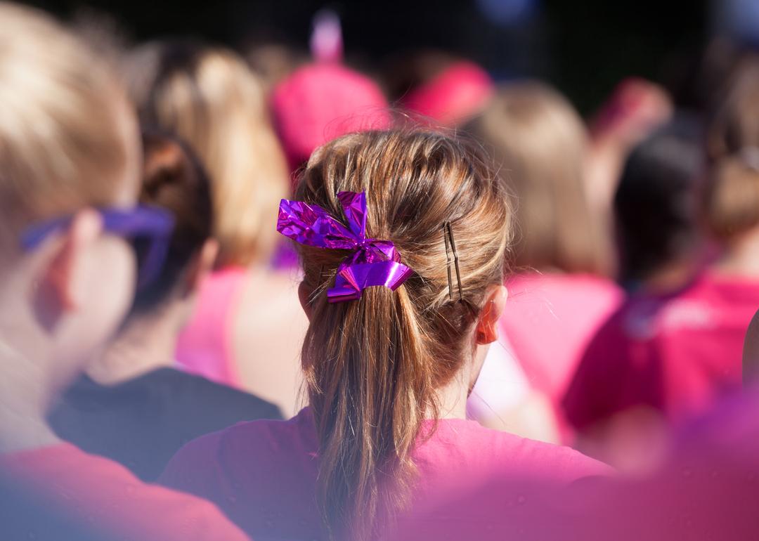 Woman, photographed from behind, stands in a crowd wearing a pink pony tail with a pink ribbon as a symbol for fighting cancer. She is surrounded by ladies wearing pink, all raising money to fight cancer
