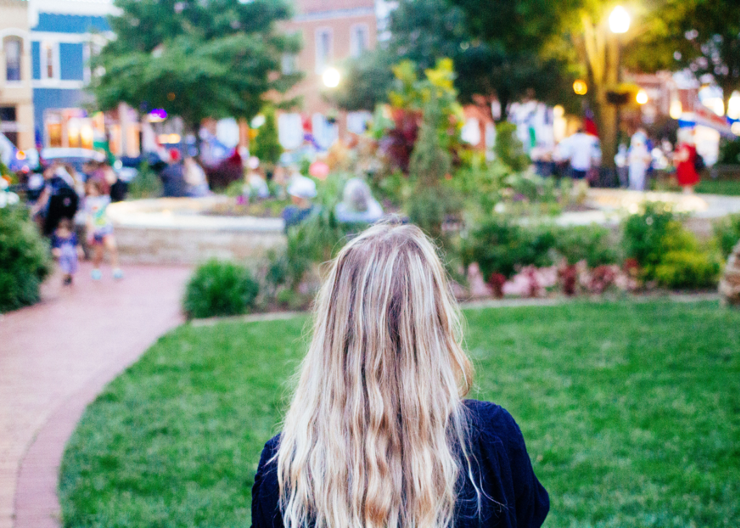 Blonde woman standing in town square with lush green grass and lots of people.