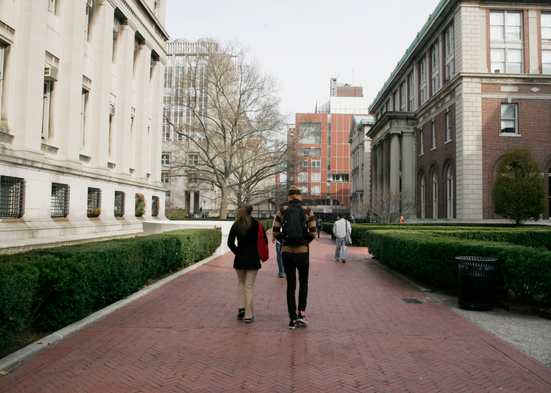Students with backpacks walk with their backs to the camera on a college campus, surrounded by buildings.