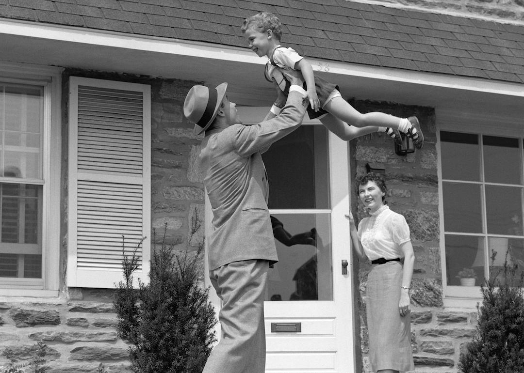 A 1950s father coming home and lifting his son in the air while his wife looks on from the doorway.