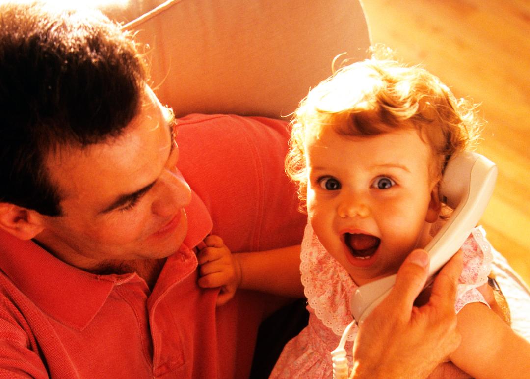 Father holds infant who's looking at the camera while listening to someone on a corded telephone in the 1990s.