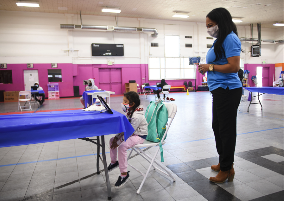 A teacher in a mask helping a student on a computer during COVID-19.