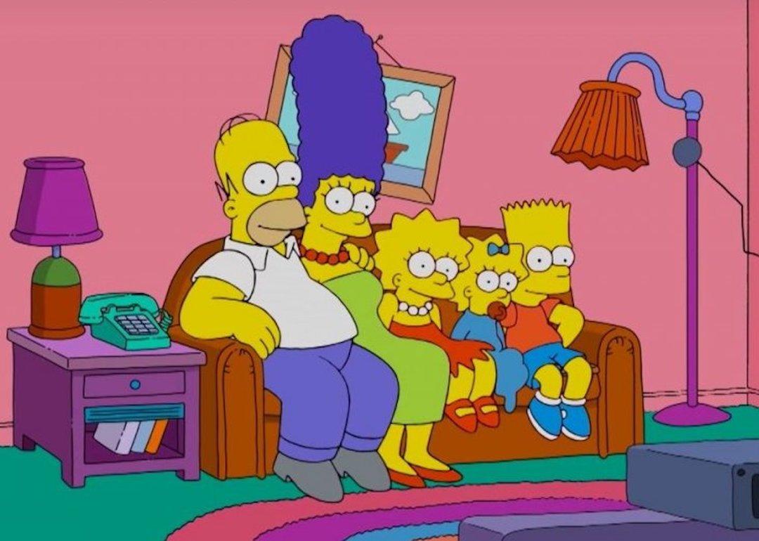 Homer, Marge, Lisa, Maggie, and Bart Simpson sit on their couch watching TV on "The Simpsons"