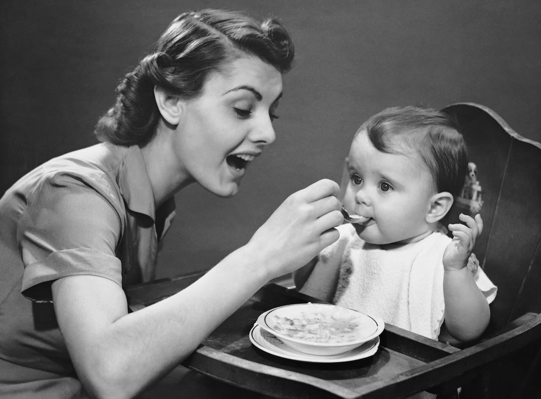 Woman feeding baby in a high chair in the 1950s.