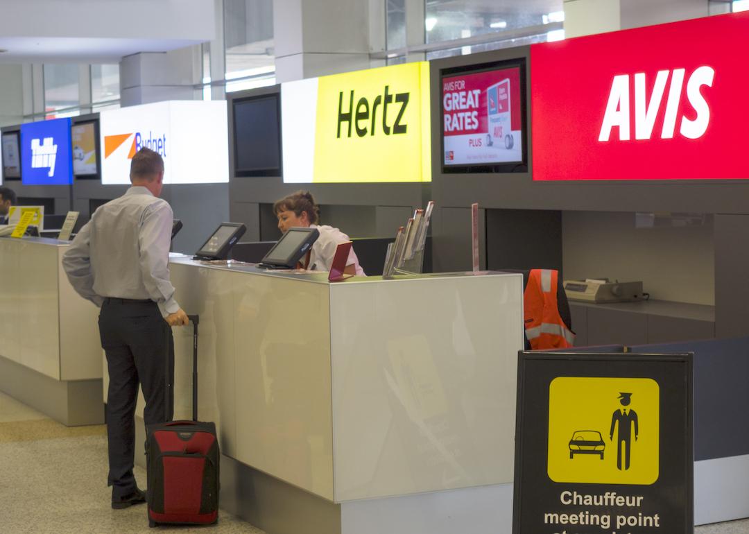 Man at airport rental car counter talks to Avis clerk, who's next to Hertz, Budget, and Thrifty counters.