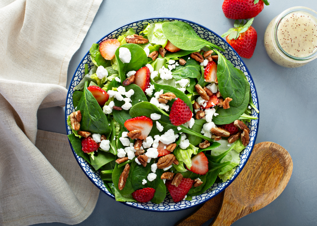 Strawberry, goat cheese, and walnut salad in a bowl, sits next to wooden spoon and dressing.