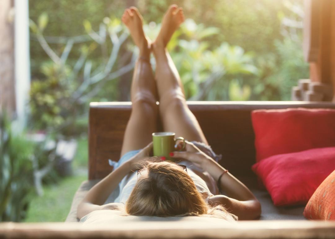 Woman drinking out of a mug while lying on a couch on her porch.