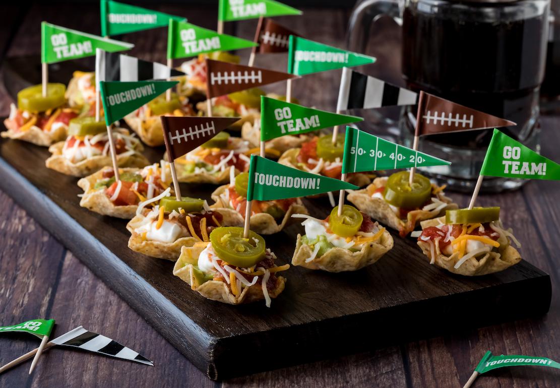 A close up view of a wooden platter of layered dip appetizers topped with jalapeños ready for a football viewing party.
