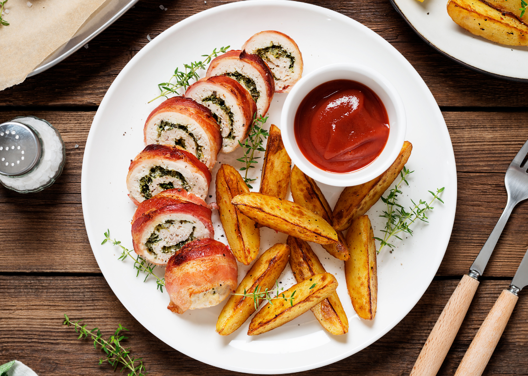 Bacon-wrapped chicken rolls on a white plate with homemade fries and ketchup on the side.