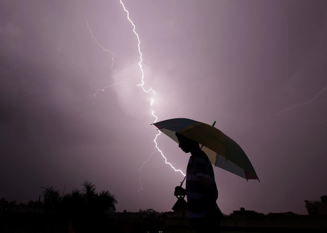 A pedestrian walks with an umbrella as lightning strikes during an evening thunderstorm